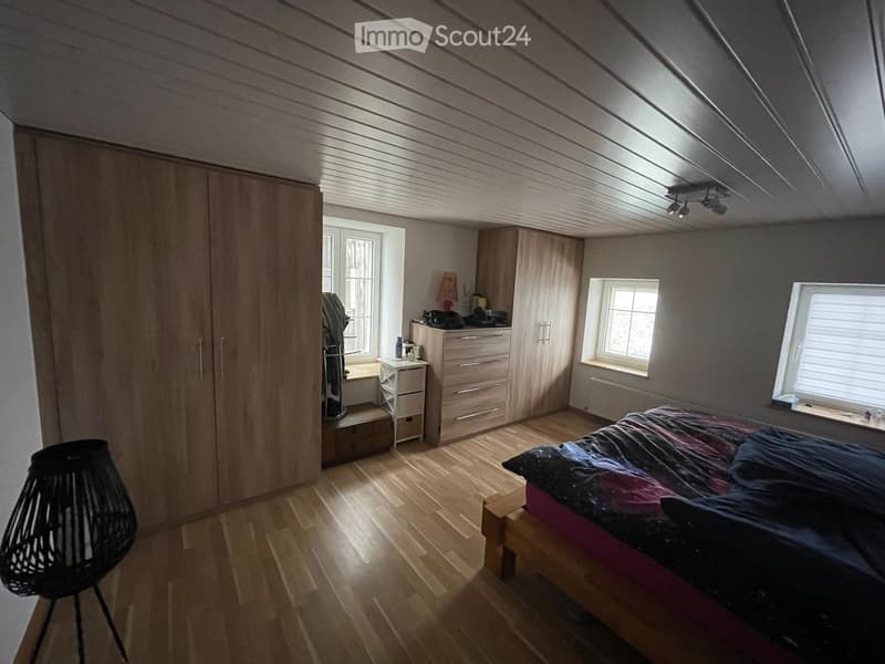 Bedroom with wooden floor, slanted ceiling, large windows, bed with colorful bedding, wardrobe, small table, and chair.