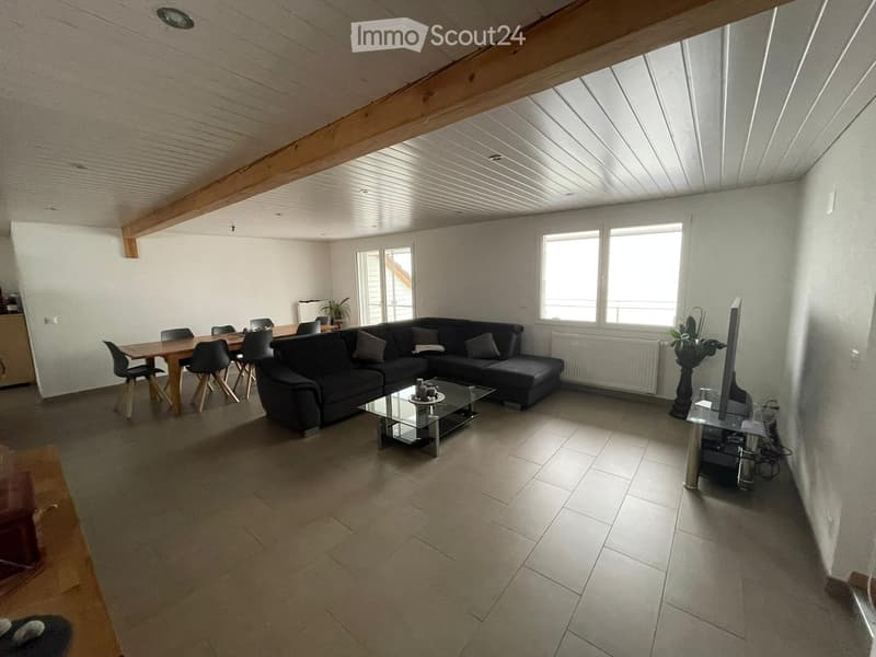 Bright living room with a black sectional sofa, glass coffee table, dining area with wooden table and chairs, and a TV stand with a flat-screen TV. The room features exposed wooden beams and large windows allowing natural light.