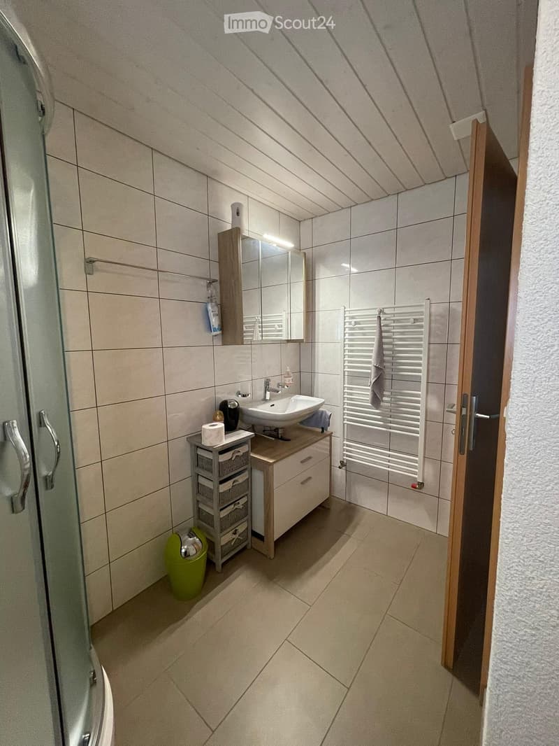 Modern bathroom with tiled walls and floor, a sink with a mirror cabinet, a towel rack, a small storage unit, and a green trash bin.