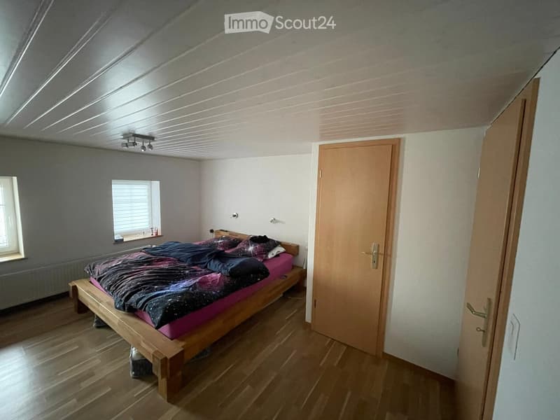 Bedroom with wooden floor, slanted ceiling, bed with colorful bedding, and a door leading to another room.
