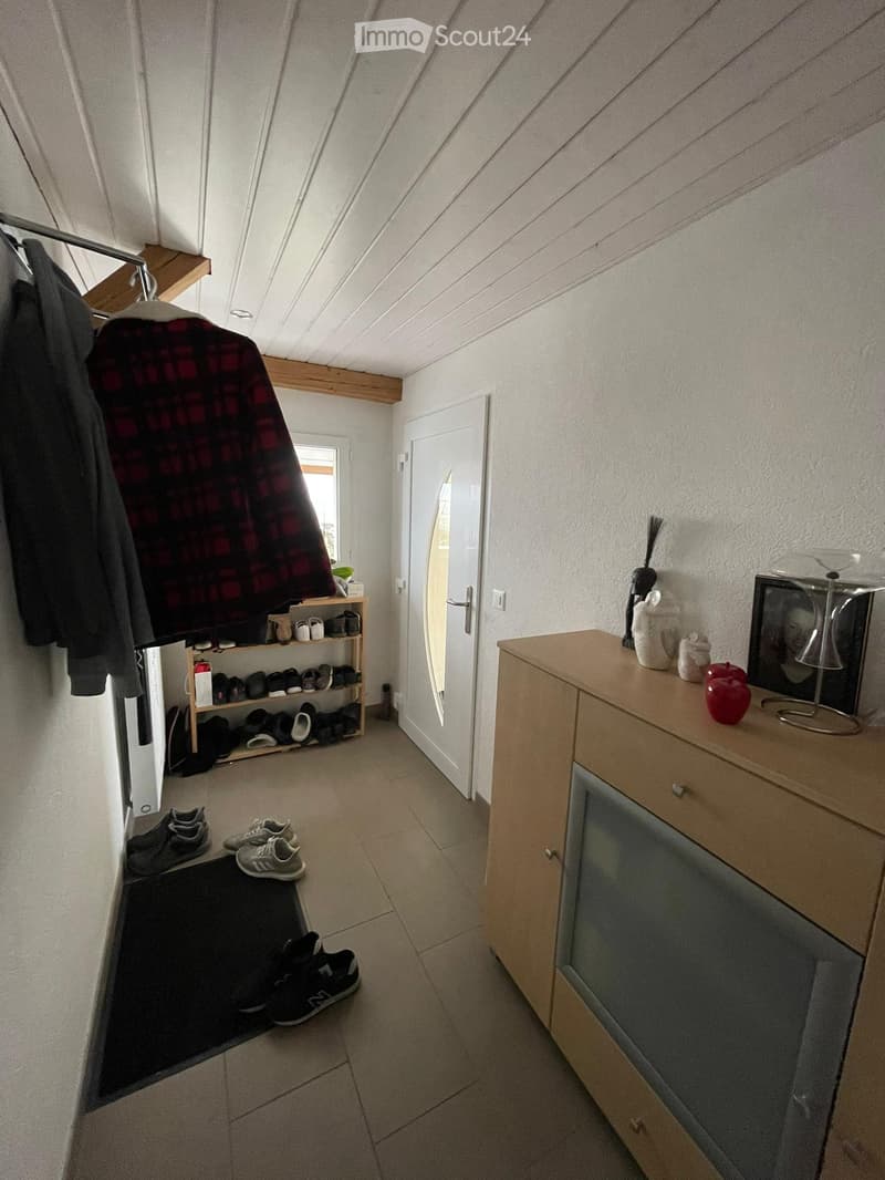 A narrow hallway with a slanted ceiling, tiled floor, and a coat rack with jackets and shoes. A small wooden shelf holds shoes and a broom, while a wooden cabinet with a mirror and decorative items stands on the right side.