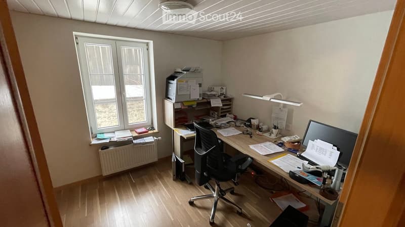 Office space with a wooden floor, a large window, a desk with a computer screen, a chair, a printer, and a lamp.