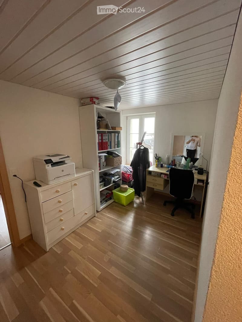 Attic room with wooden flooring, slanted ceiling, large window, desk, chair, cabinet, shelf, and mirror.