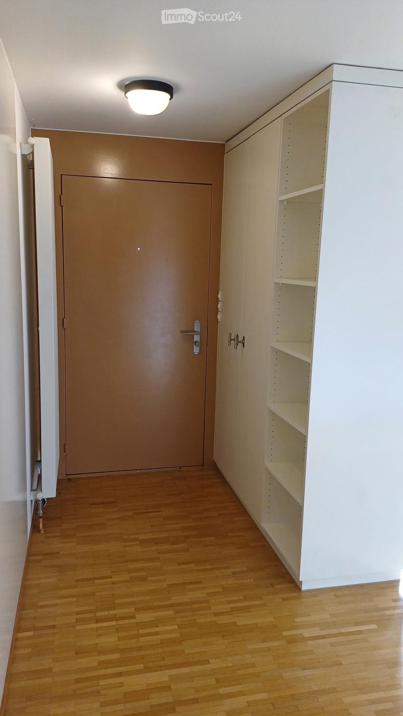 Empty hallway with wooden flooring, white walls, a closed brown door, and built-in white shelving unit with empty shelves.