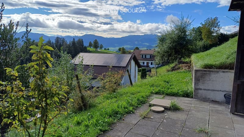 Garden area with a view of mountains, houses, and greenery, featuring a paved patio and small plants.