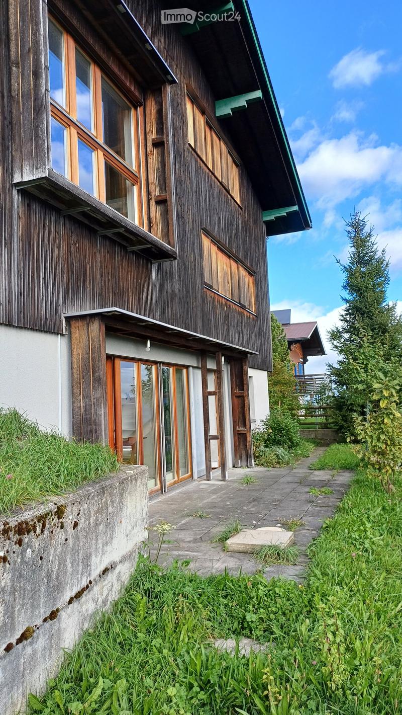 Two-story wooden house with large windows, green roof trim, and a concrete pathway leading to the entrance.
