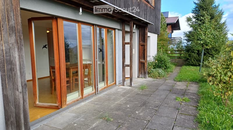 Modern house exterior with large sliding glass doors leading to a wooden-floored interior, visible dining area with chairs and table, surrounded by greenery and a paved patio.