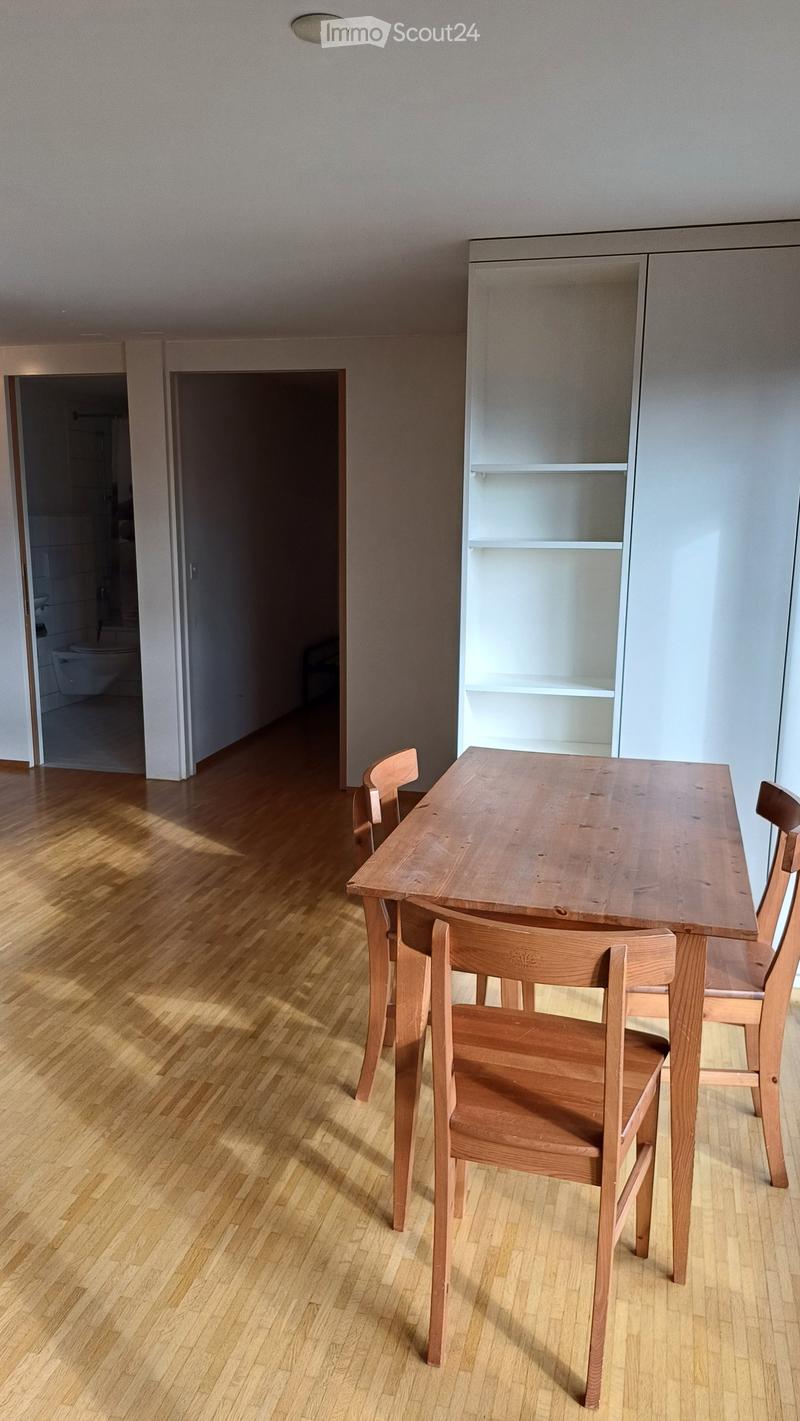 A modern, minimalist dining area with a wooden table and four matching chairs, a built-in white cabinet, and a view into an adjacent bathroom.