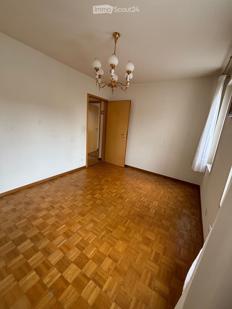 Empty room with wooden herringbone flooring, white walls, and a chandelier hanging from the ceiling. Two open doors lead to adjacent spaces.