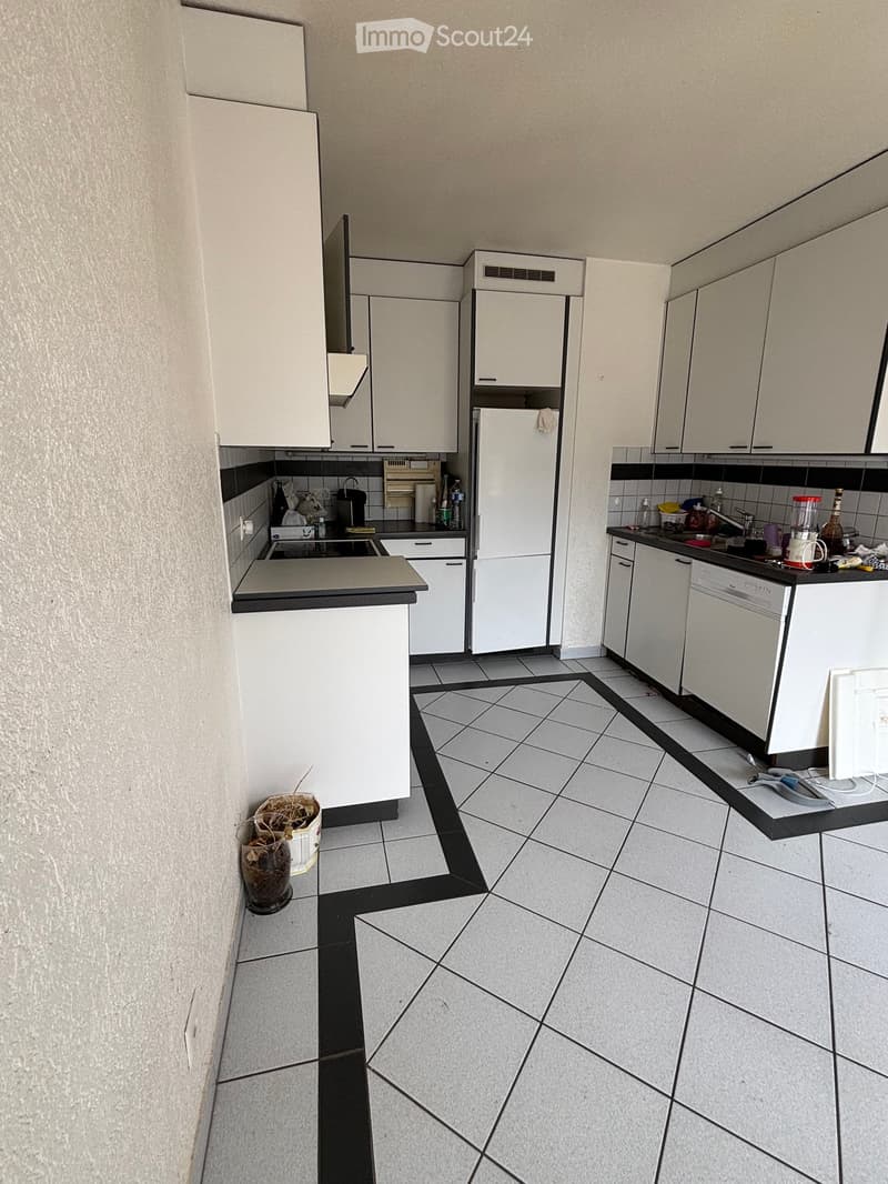 Modern kitchen with white cabinets, tiled floor, and various appliances including a refrigerator, dishwasher, and oven. Countertops are visible with a sink and faucet, and there are several small items and containers on the counters.