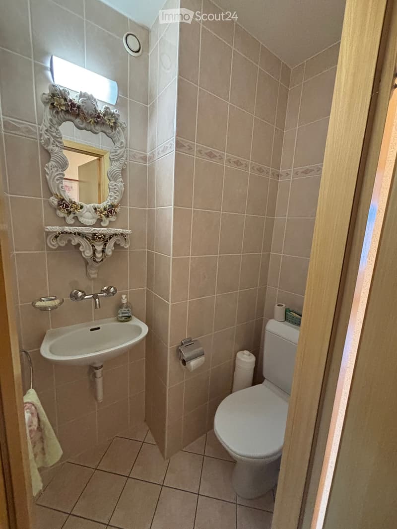 Small bathroom with beige tiled walls and floor, featuring a sink with a decorative mirror, a toilet, and a towel hanging on the side.