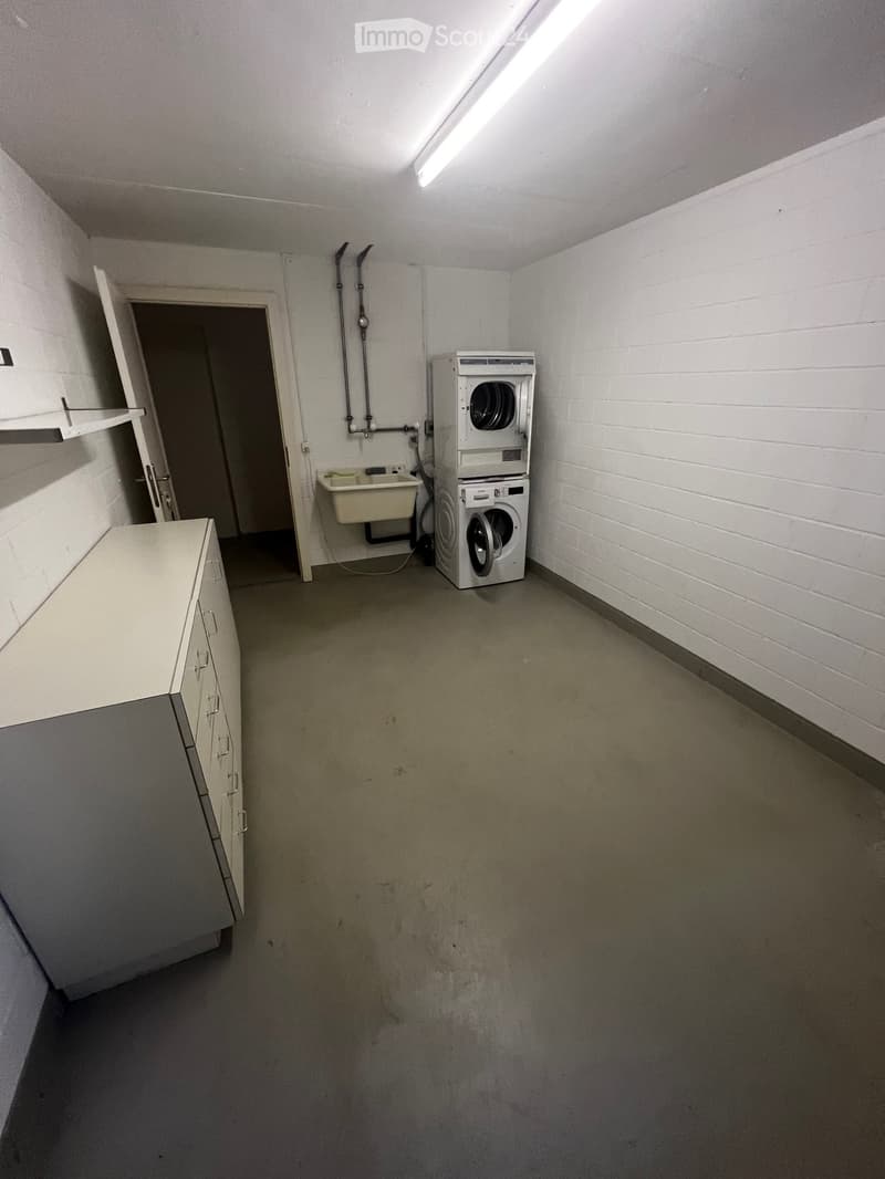 A small utility room with a washing machine, a sink, and a cabinet, featuring white brick walls and a concrete floor.