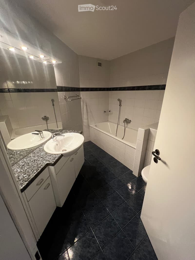 Modern bathroom with two sinks, a bathtub, and a large mirror with built-in lighting. Dark tiled floor and white tiled walls.