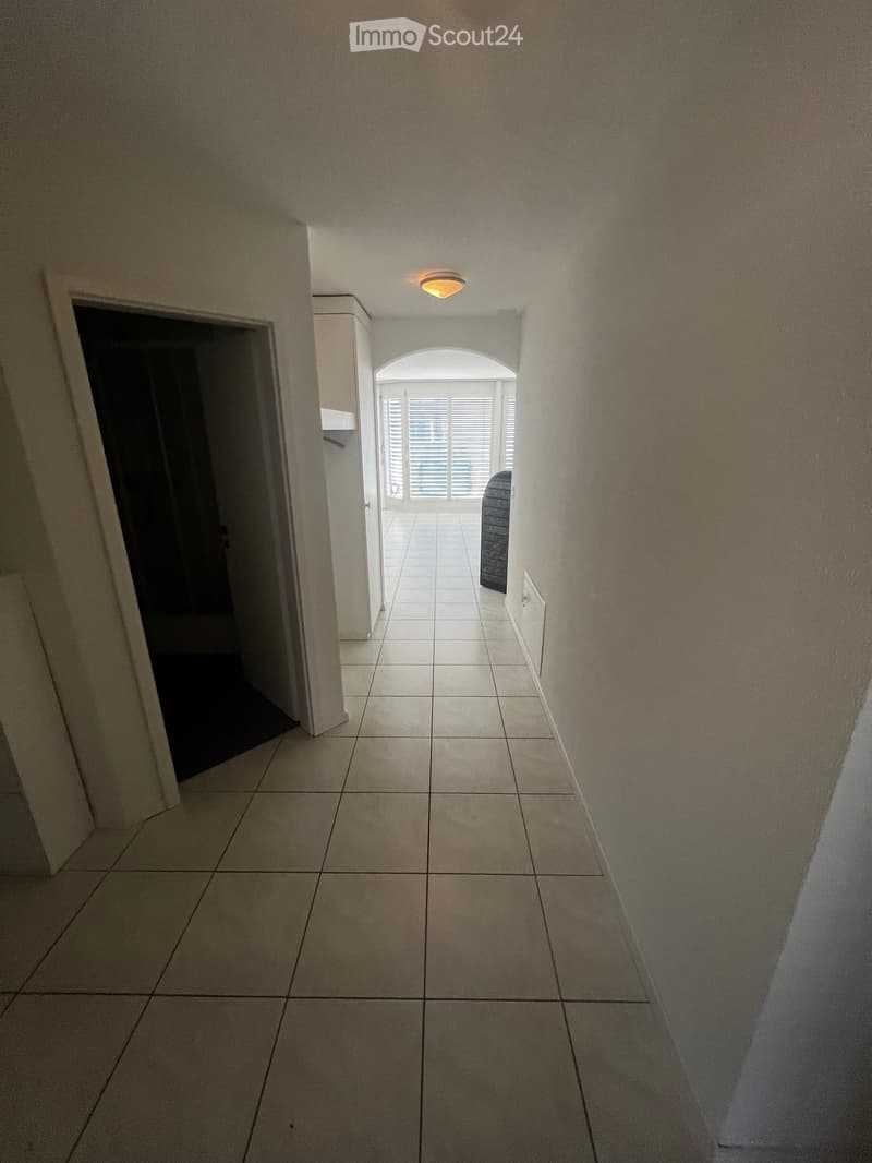 A clean, tiled hallway with beige walls, a ceiling light fixture, and an arched opening leading to a room with white blinds and a visible piece of furniture.
