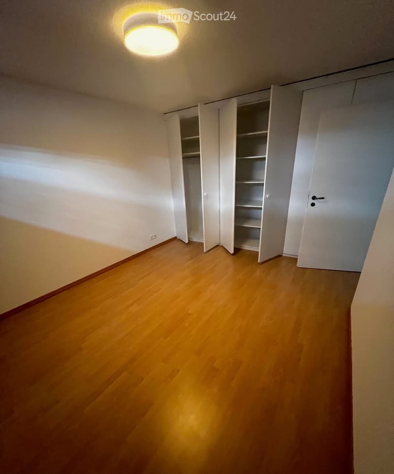 Empty room with wooden flooring, white walls, and a built-in closet with empty shelves. A round ceiling light fixture is visible.