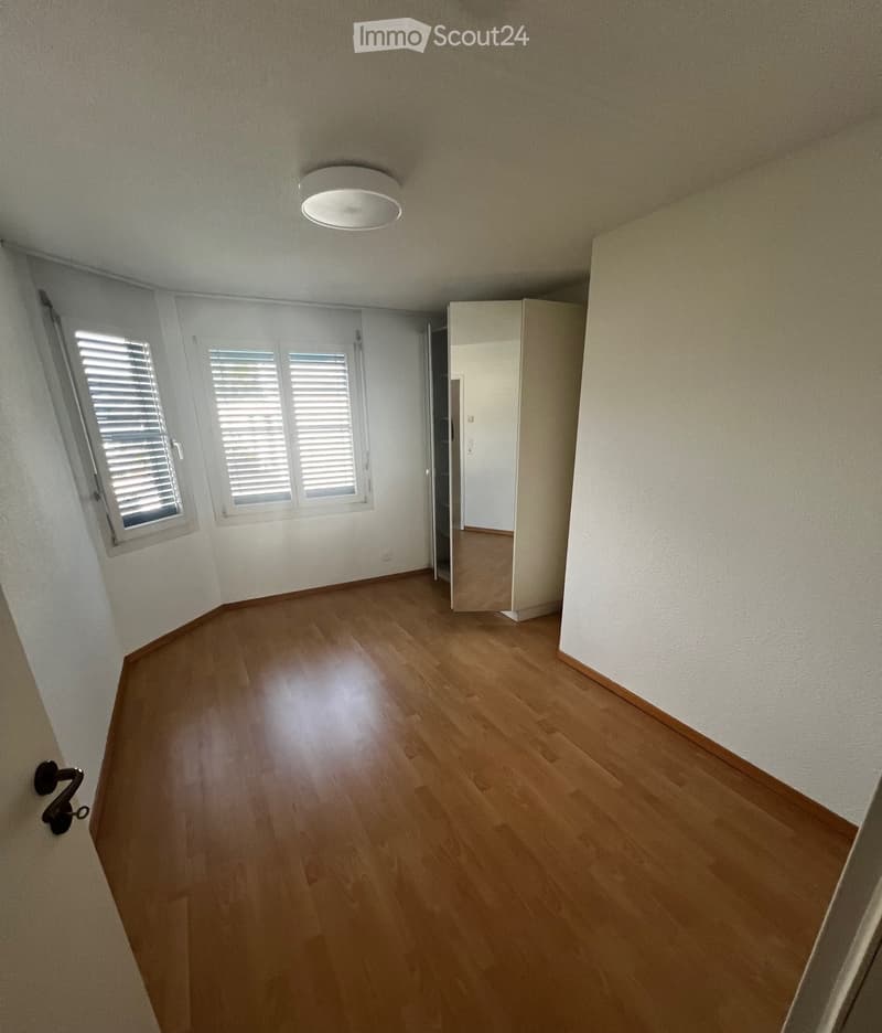 Empty room with wooden flooring, white walls, and a single round ceiling light. Windows with blinds are visible on one wall, and a partially open door leads to another room.
