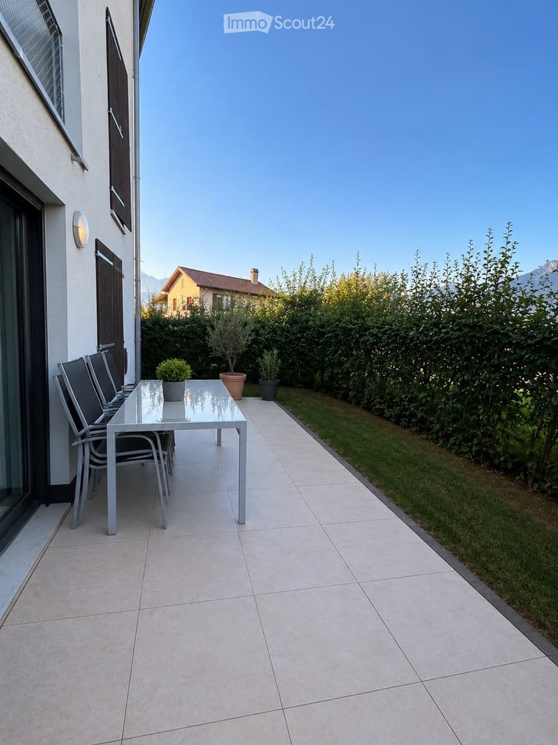 A modern outdoor patio with tiled flooring, a glass dining table, and four matching chairs. Potted plants are placed near the edge, and the area is bordered by a neatly trimmed hedge and a lawn.