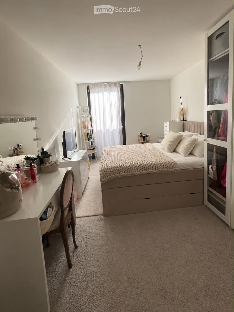 A well-lit bedroom with a bed, a chair, a mirror, a desk, a wardrobe, and a curtain covering a window. The room features a cozy, neutral color palette with a carpeted floor.