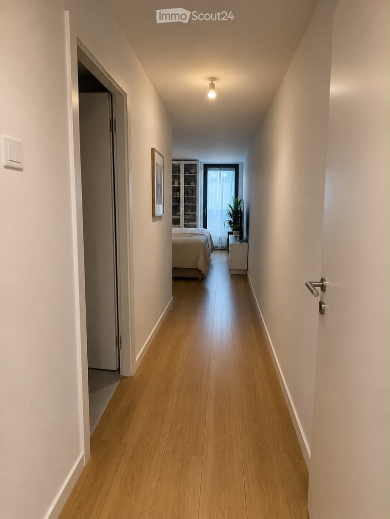 A narrow hallway with light wooden flooring, white walls, and a single ceiling light. An open door on the left leads to a room with a bed and a small cabinet, while a framed picture hangs on the wall.