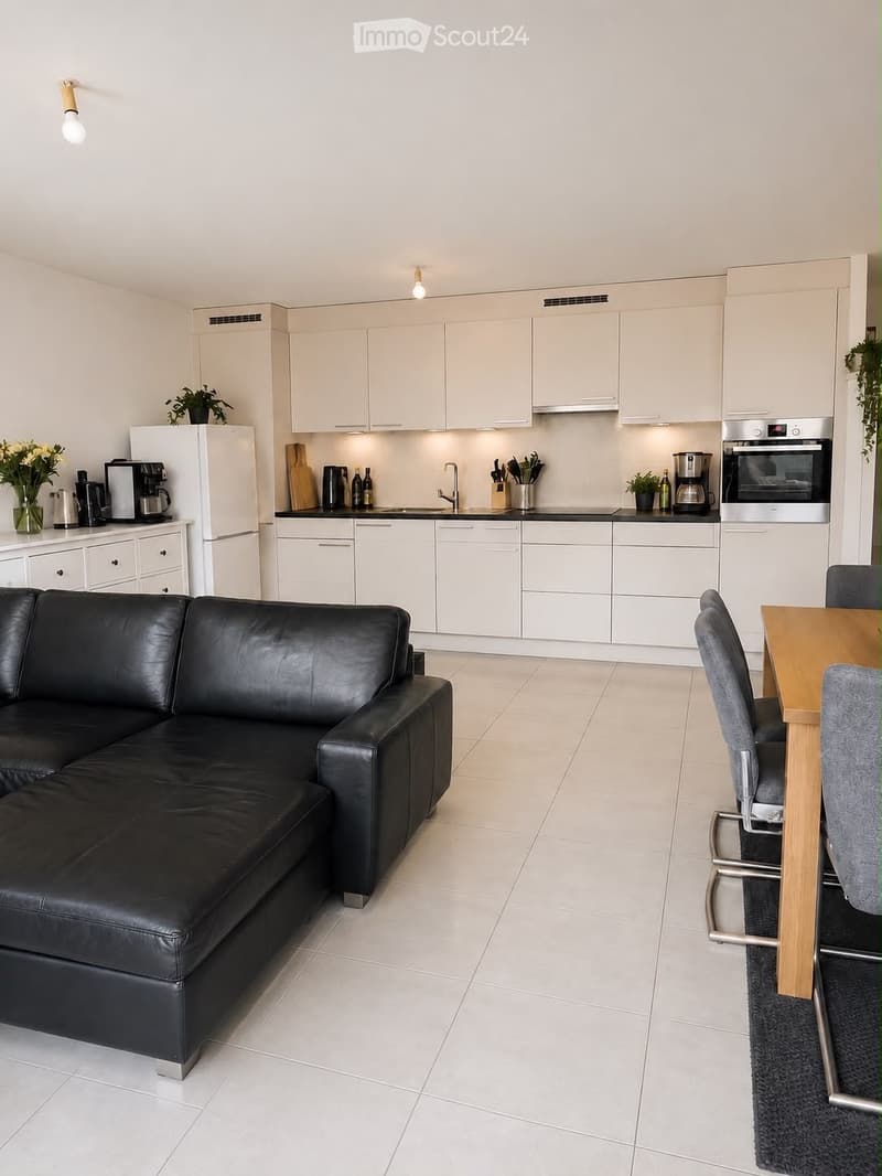 Open-plan living and dining area with a black leather sofa, a wooden dining table with chairs, and a modern kitchen featuring white cabinets, stainless steel appliances, and a countertop with various items.