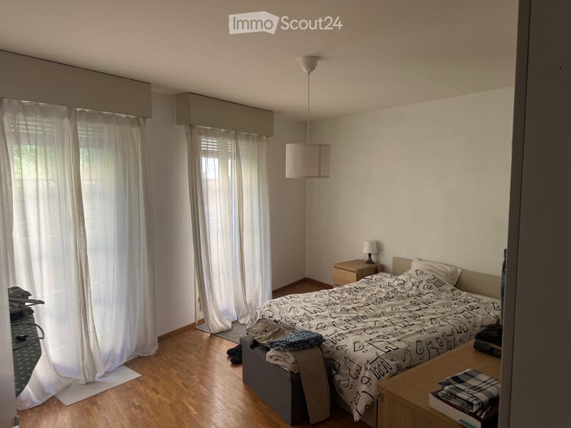 A modern bedroom with a wooden floor, a bed with a patterned duvet, a nightstand with a lamp, and large windows with white curtains.