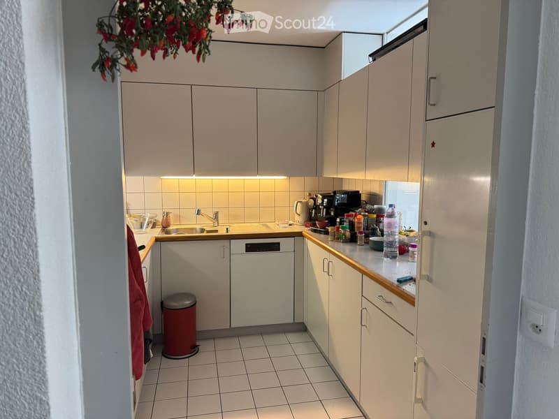 Modern kitchen with white cabinets, tiled floor, and various appliances including a coffee maker, refrigerator, and oven. Countertop has a bottle, cup, and other small items.