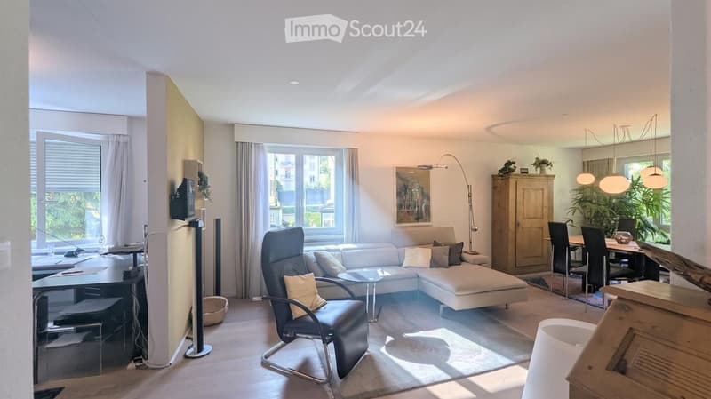 Bright living room with a white sectional sofa, coffee table, armchair, dining area with table and chairs, large windows with curtains, and a floor lamp.
