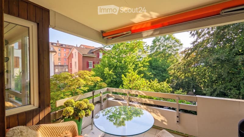 Balcony with a glass table, a chair, and a vase of flowers, overlooking a green, tree-filled area with neighboring houses.