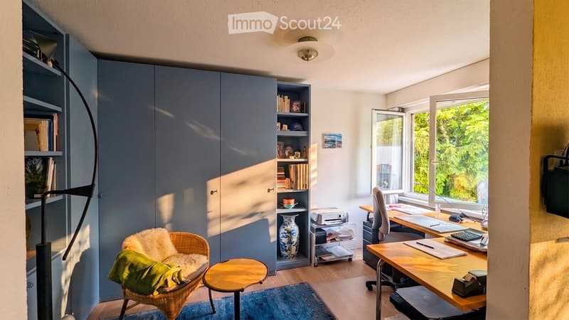 A modern, well-lit room with a mix of furniture including a wicker chair, a small wooden stool, a large wooden desk, a cabinet, and shelves filled with books and decorative items. The room features large windows with a view of greenery outside.