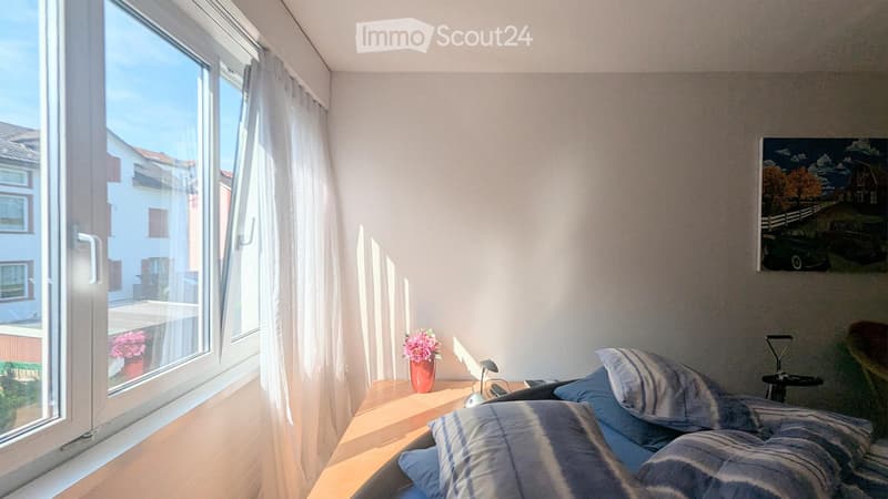 Cozy bedroom with a bed, nightstand, lamp, vase with flowers, and a painting on the wall. Large window with white curtains offers natural light and a view of the street.