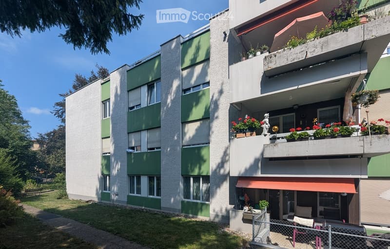 Modern multi-story apartment building with green and gray facade, large windows, balconies with potted plants and flowers, and a small fenced garden area in front.