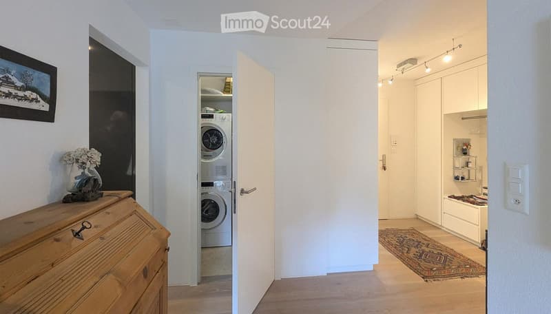 A well-lit hallway with wooden flooring, leading to a laundry area with a washing machine and dryer, and a small bathroom with a bathtub and shelves.