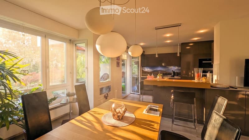 Bright, modern open-plan living and dining area with large windows, a wooden dining table with chairs, a kitchen island, pendant lights, and decorative items.