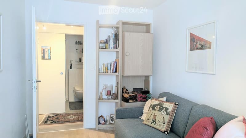 A modern living room with a grey sofa, a wooden bookshelf filled with books and decorative items, a framed picture on the wall, and an open door leading to a small bathroom.