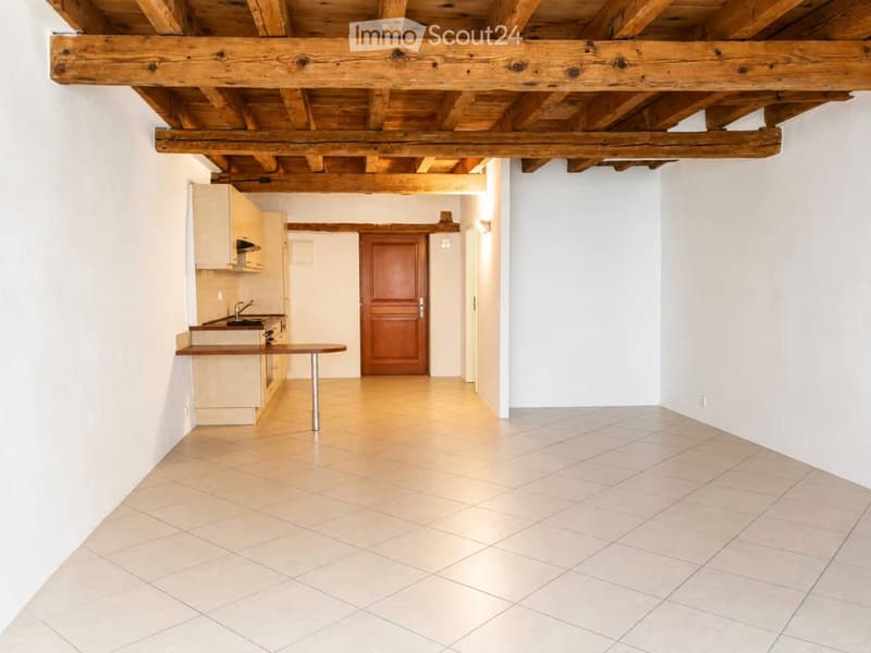 Empty room with exposed wooden beams on the ceiling, tiled floor, and a small kitchen area with cabinets and a sink visible in the background.