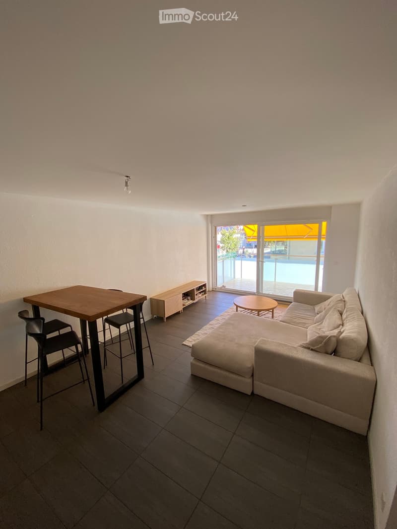 Modern living room with a grey sectional sofa, coffee table, dining table with chairs, and a small cabinet. The room features large windows with a view of a swimming pool and outdoor area.