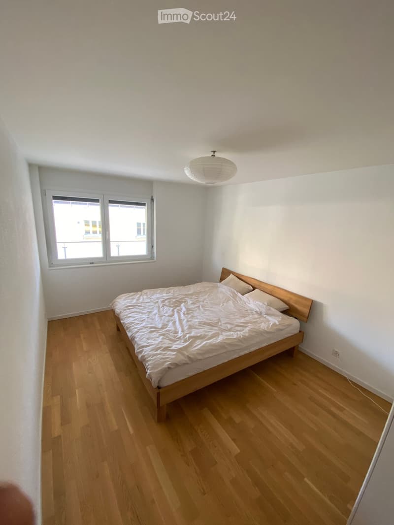 A small, unfurnished bedroom with a wooden bed, white bedding, and a single window letting in natural light.