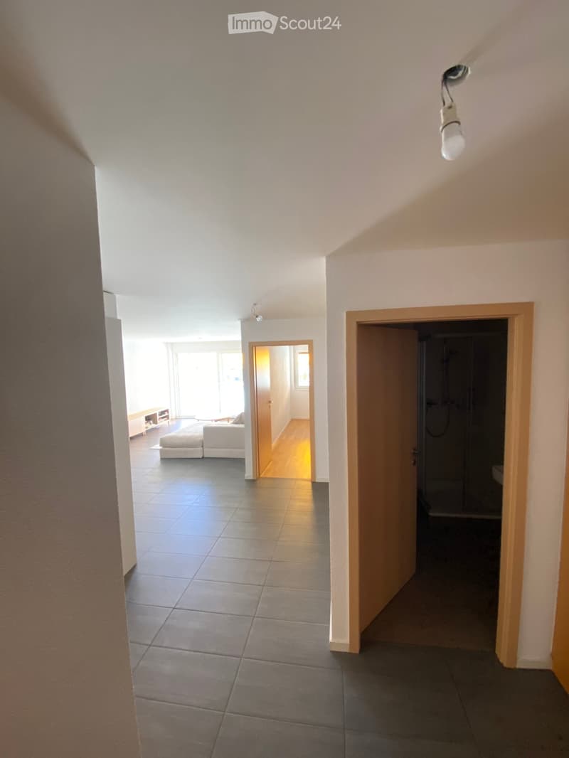 Bright hallway with tiled floor, leading to a living area with white seating and a small table, and a bathroom with a shower stall.