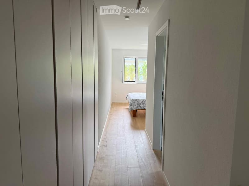 A narrow hallway with light wooden flooring, white walls, and a visible bedroom at the end with a bed and patterned bedding.