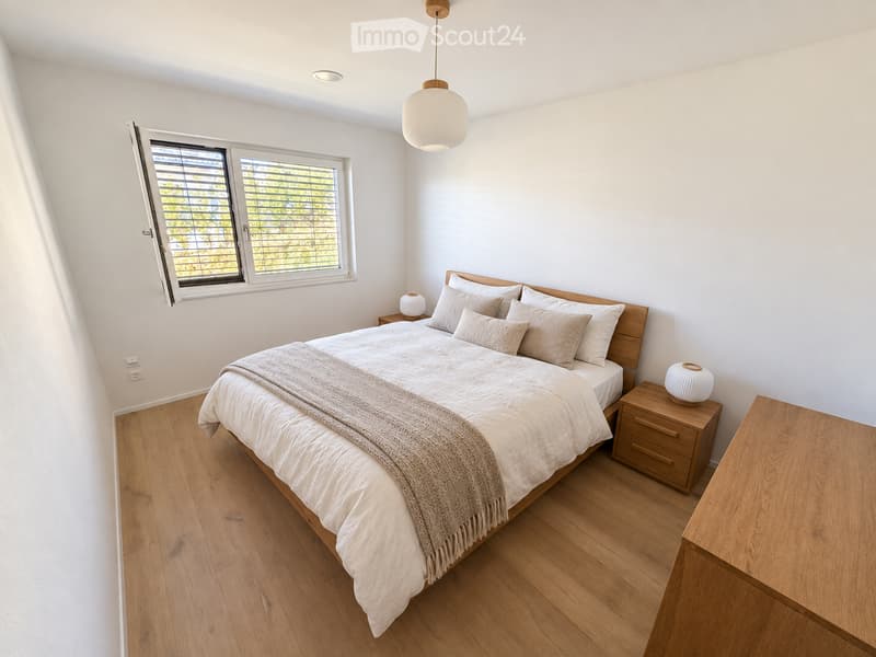 Modern bedroom with a large bed, wooden nightstand, and pendant lighting. The room features light-colored bedding, a wooden floor, and large windows with blinds.