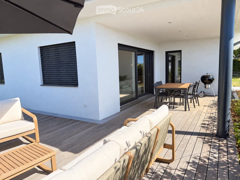 Modern outdoor patio with wooden decking, white walls, large sliding glass doors, and a covered seating area with a sofa, chairs, and a table. A grill is positioned near the edge of the patio.