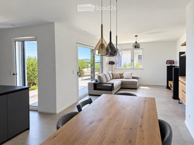 Modern living and dining area with a grey sectional sofa, wooden dining table, pendant lights, and large windows offering a view of greenery.