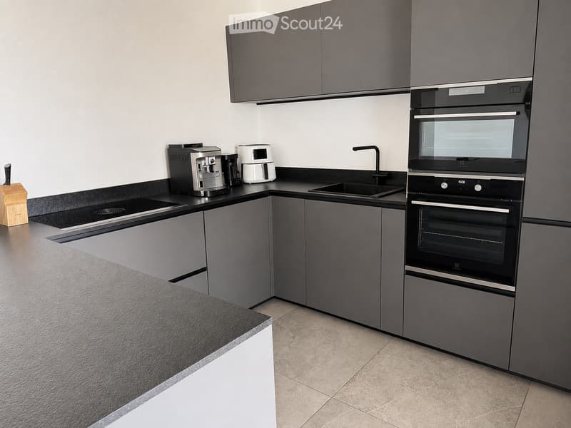 Modern kitchen with grey cabinets, black countertops, and stainless steel appliances including a coffee maker, toaster, and built-in oven.