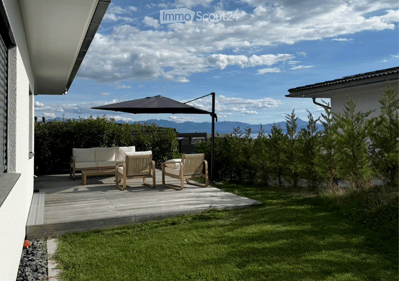 A modern outdoor patio with a wooden deck, white sofa, two wicker chairs, and a large umbrella, surrounded by greenery and a scenic mountain view.