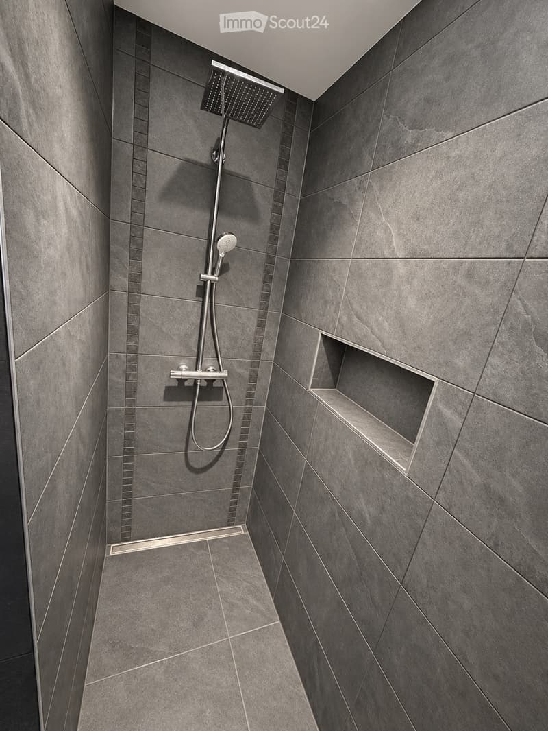 Modern bathroom shower area with grey tiled walls and floor, featuring a handheld showerhead, a fixed showerhead, a built-in shelf, and a sleek chrome shower rail.