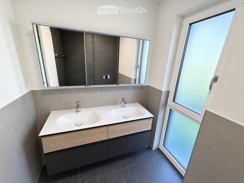Modern bathroom with a double sink, sleek wooden vanity, large mirror, and a large window providing natural light.