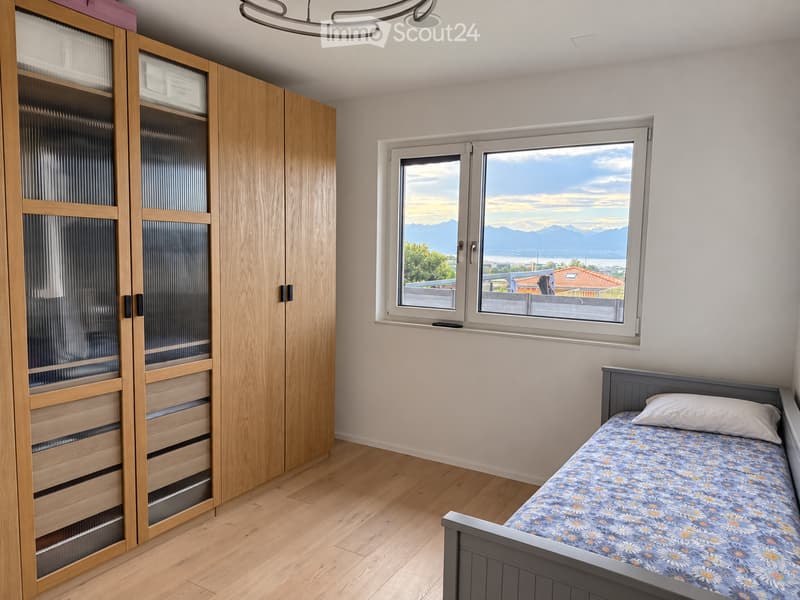 Bright bedroom with wooden floor, large window offering a scenic mountain view, single bed with floral bedding, and a tall wooden wardrobe with mirrored doors.