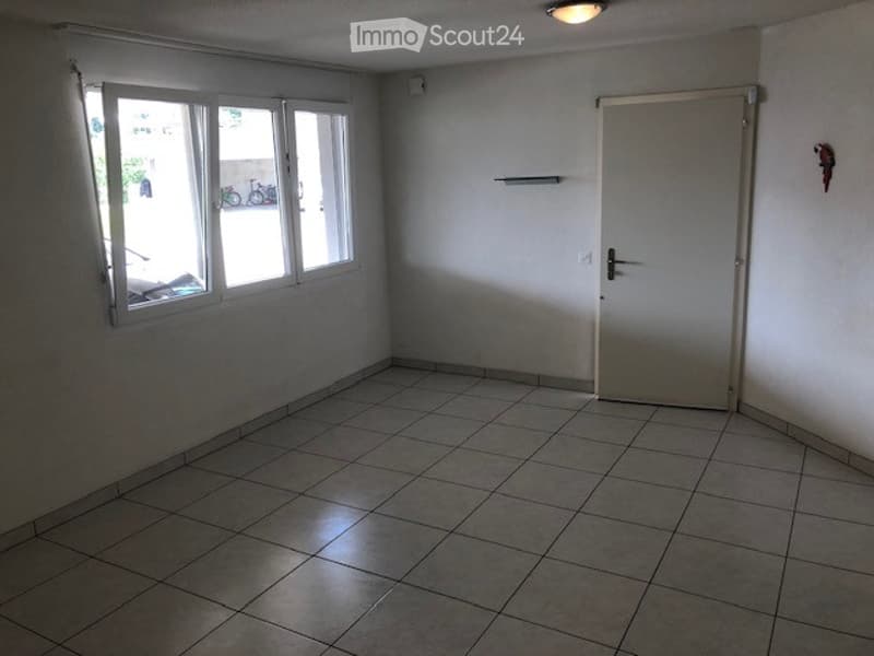Empty room with tiled floor, white walls, large windows, and a closed door.