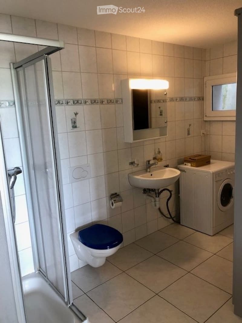 Modern bathroom with white tiled walls and floor, featuring a toilet, sink, mirror, shower, and a private washing machine.