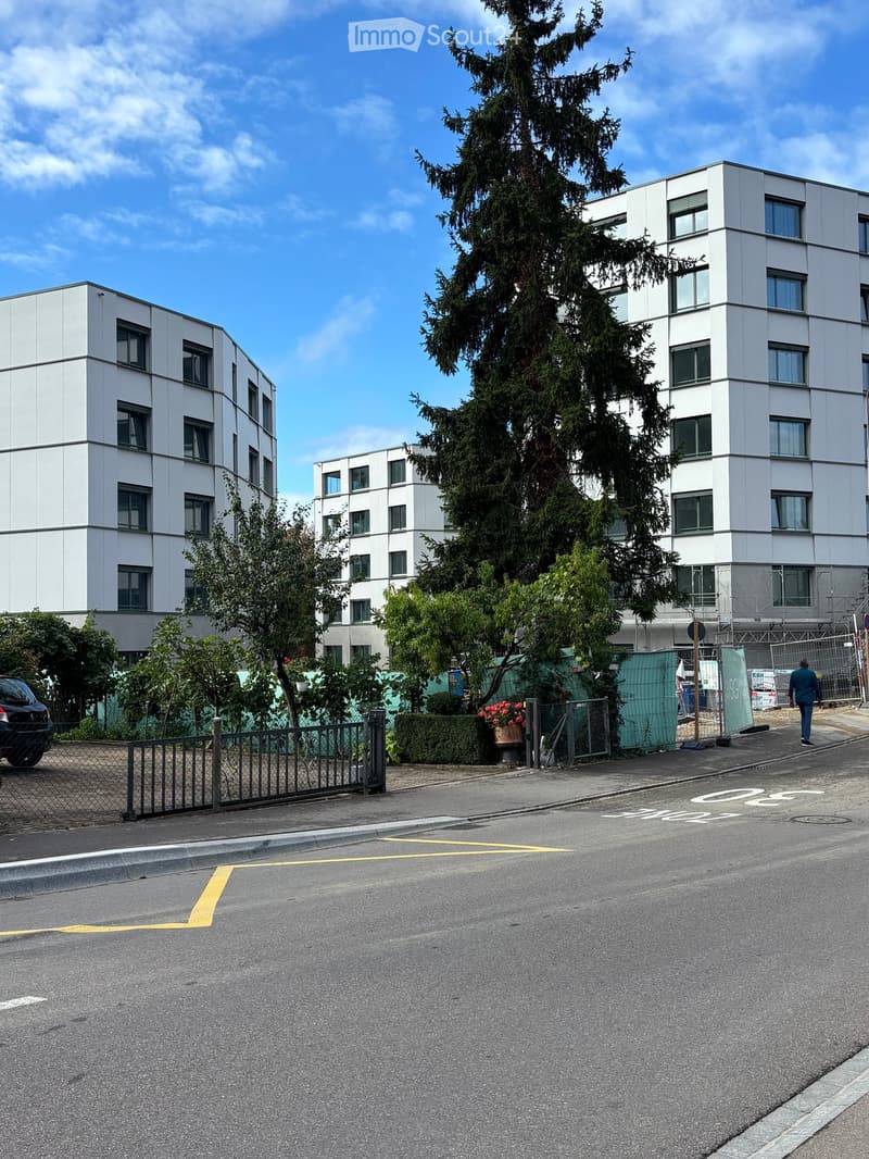 Modern multi-story residential building with white facade and large windows, surrounded by trees and greenery. A fenced area with a parked car is visible in the foreground.
